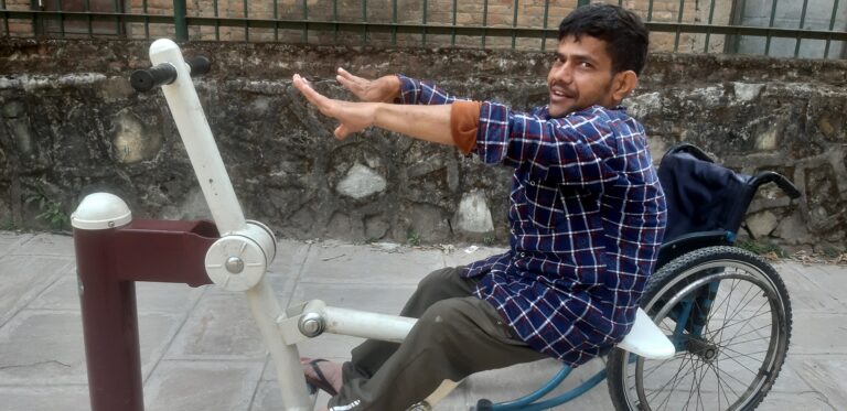 Person in a plaid shirt sits on a handcycle, reaching forward. Stone wall and metal fence in the background.