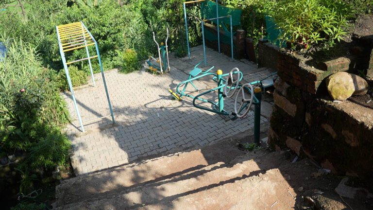 Outdoor fitness area with exercise equipment on a paved surface surrounded by greenery. Stone steps lead down to the area.