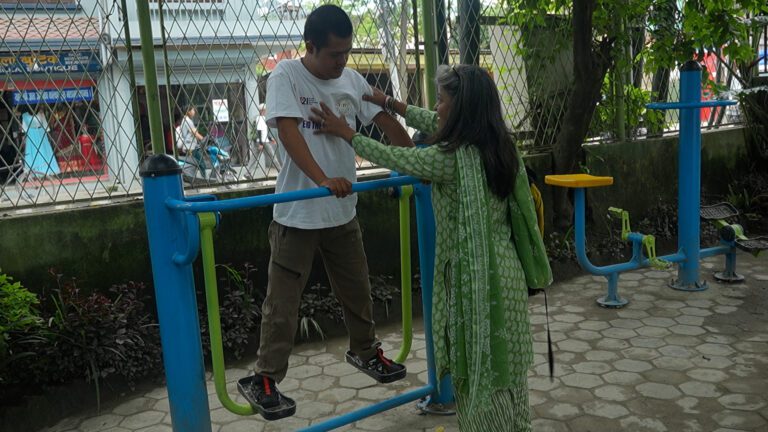 A person helps another holding onto parallel bars in an outdoor exercise area with fitness equipment.
