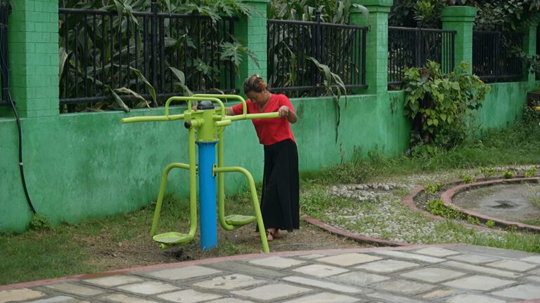A person in a red shirt uses an outdoor exercise machine next to a green fence and foliage.