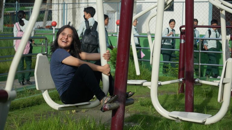 A person smiling while sitting on playground equipment. Schoolchildren in uniforms walk in the background.