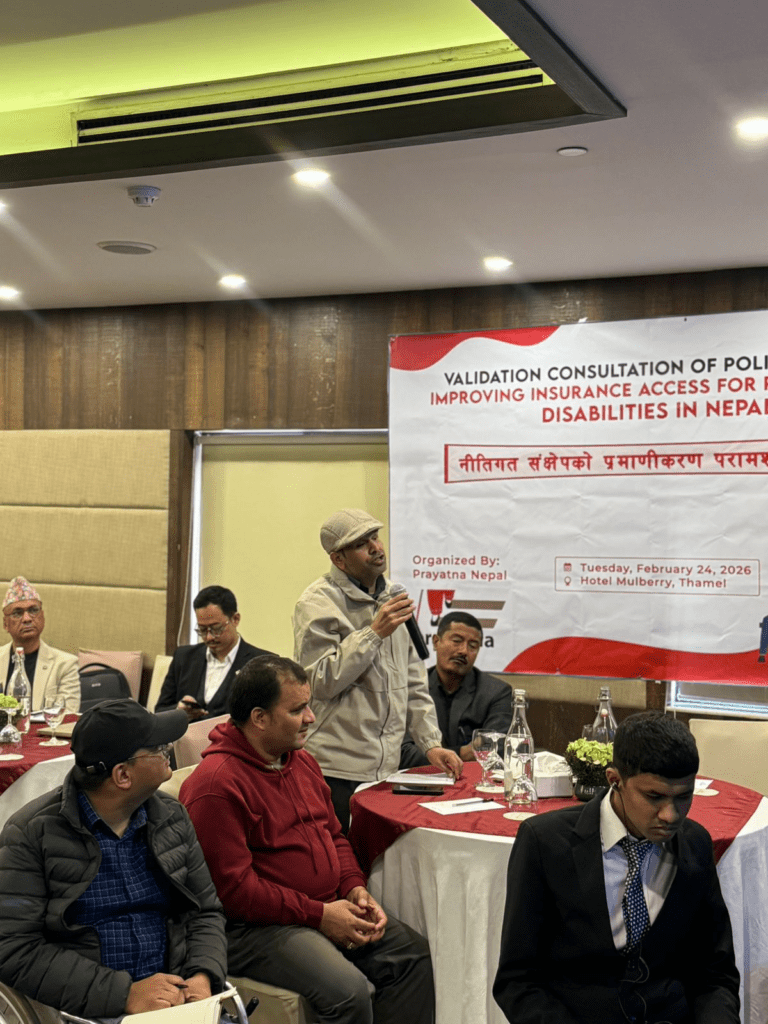 A man stands and speaks into a microphone during a validation consultation meeting on insurance access for people with disabilities in Nepal. Other participants are seated around him.