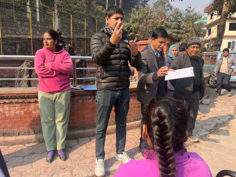 A group of people stands outdoors; one man speaks while gesturing, another holds papers, and others listen or look on.