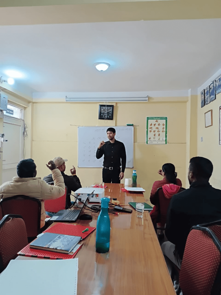 A man stands and speaks in front of a whiteboard to a group of seated people in a classroom setting, with notebooks and water bottles on the table.