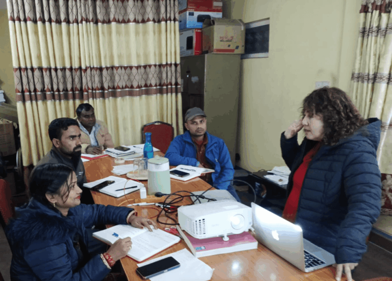 Five people are seated around a table with papers, notebooks, and a projector, while one person stands and speaks, appearing to lead a discussion or meeting in a room.