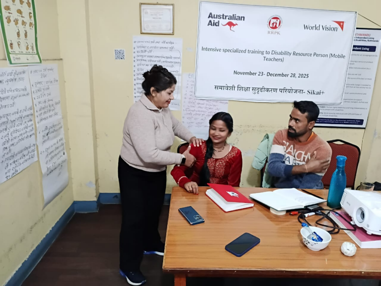 Three people sit around a table with papers and a laptop; a woman standing helps another woman adjust her clothing. A World Vision training poster is on the wall behind them.