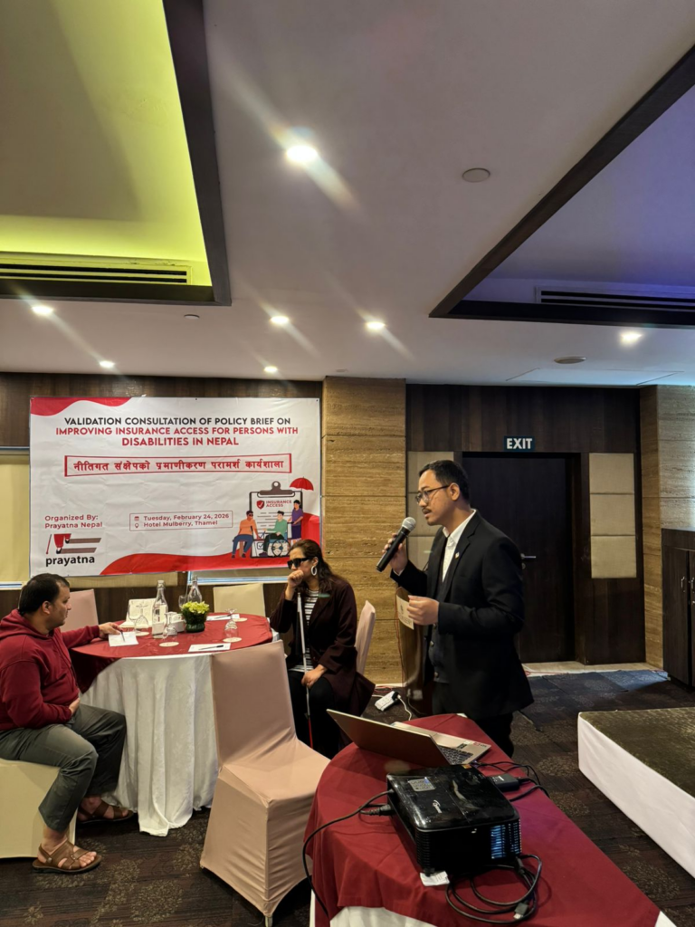 A man speaks into a microphone at a policy consultation event on improving disabilities in Nepal, with two people seated at a table and a presentation banner in the background.