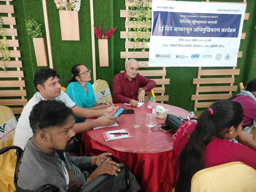 Participants seated around a round table take notes and look at a phone during a session. A participant in the foreground sits in a wheelchair, and the project banner hangs on the wall behind them.