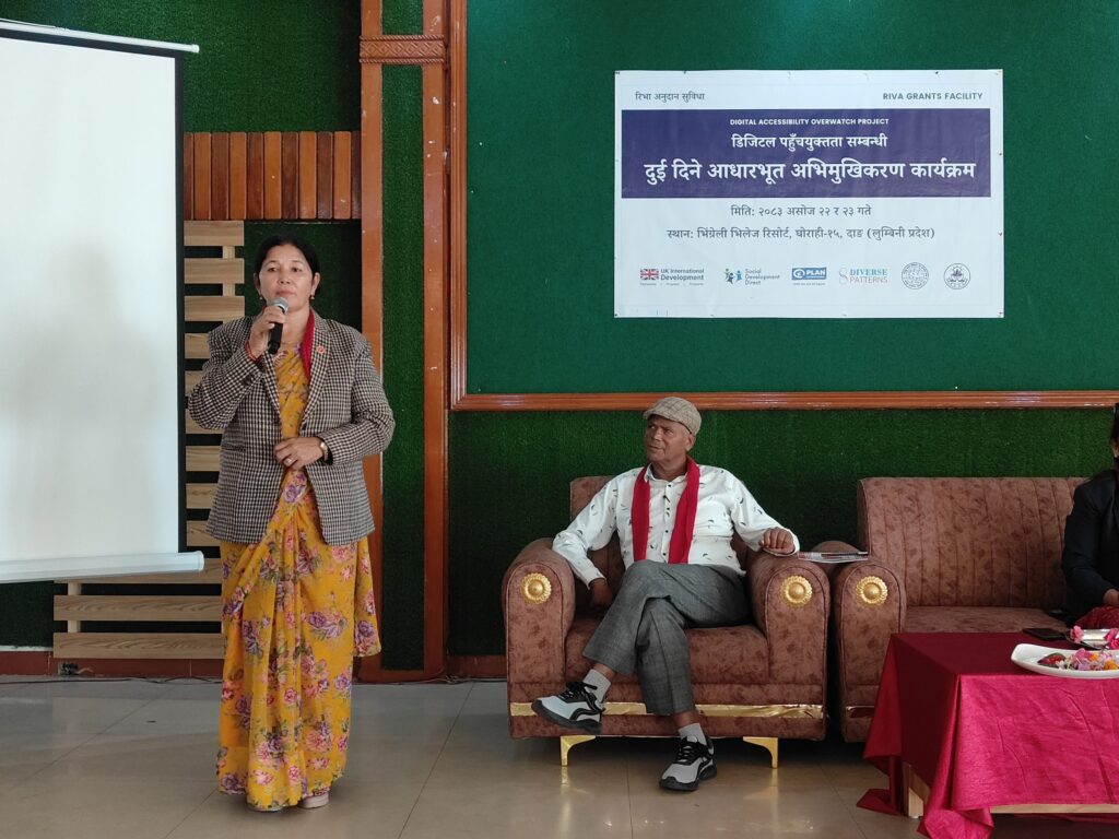A woman in a checked blazer and a yellow floral sari speaks into a microphone beside a projector screen. A man in a flat cap and a red scarf sits on a sofa behind her, under the project banner.